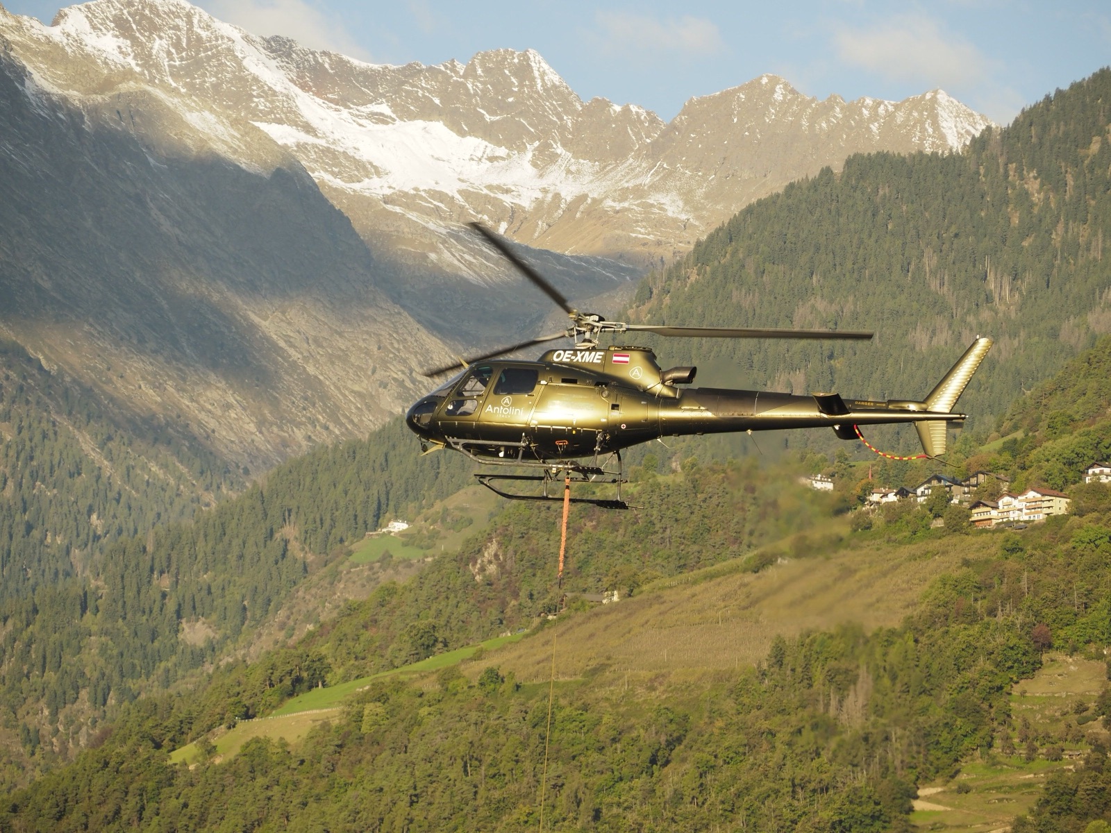 An AS350-class helicopter flying a long-line sling load through a steep mountain valley.